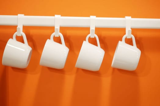 Four white mugs are neatly suspended from hooks on a vibrant orange wall.