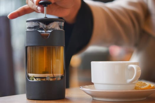 A hand is pressing down a French press filled with herbal tea beside a cup.