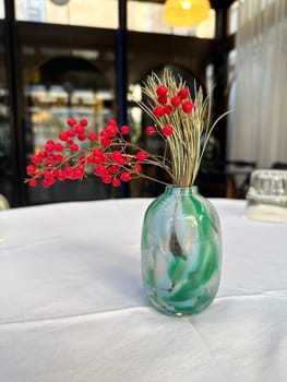 Decorative vase with red berries and pine branches on restaurant table. Minimalism, elegance, and seasonal interior decor in public spaces