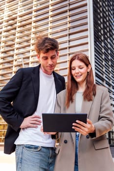 man and a woman smiling happy looking digital tablet together outdoors in front of an office building, the man is wearing a suit and the woman is wearing a jacket, concept of business and technology