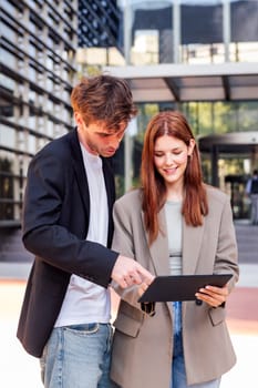 man and a woman standing outside in front of office building, the man is pointing at digital tablet in the woman hand, concept of business and technology