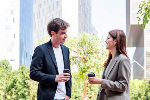 smiling man and a woman standing in a park near of office buildings talking while taking coffee cups, they are smiling and enjoying with the company, concept of business and urban lifestyle