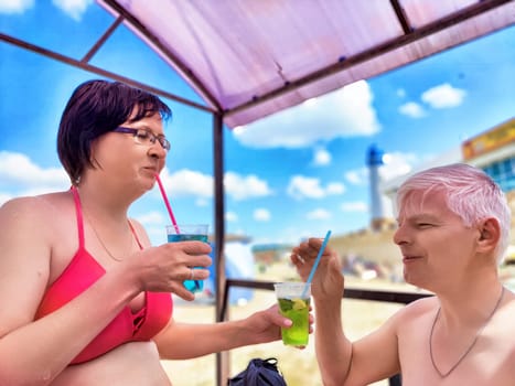 Middle-aged couple savoring cool beverages while relaxing under a cabana at the beach. They share smiles and laughter during their vacation in a warm coastal setting.