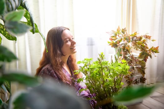 A woman with long hair smiles as she gently tends to lush indoor plants, surrounded by sunlight peeking through the curtains.