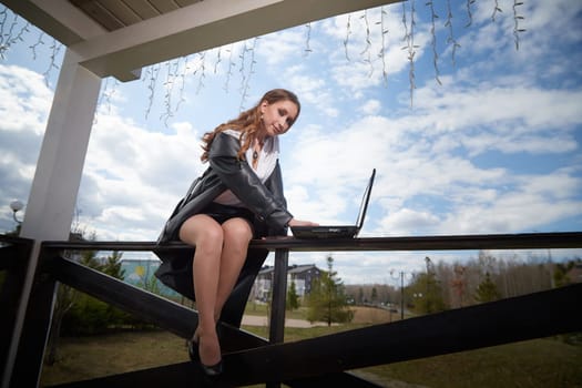 A woman sits confidently on a fence, using her laptop, with a picturesque landscape and cloudy sky in the background.