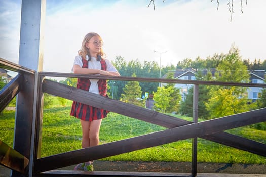 A girl in a plaid skirt leans against a wooden railing, gazing at a serene suburban park under warm sunlight.