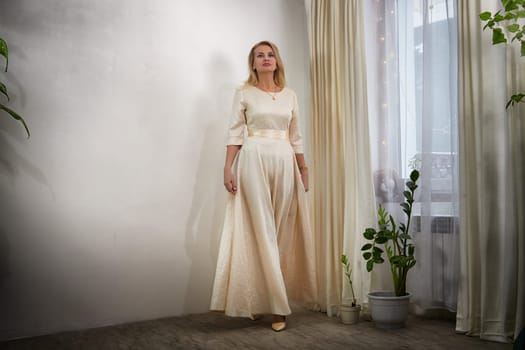A middle-aged blonde woman poses gracefully in a chic outfit during a cozy lifestyle photo shoot at home.