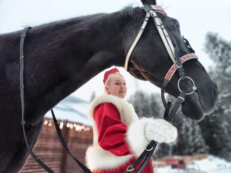 A beautiful Russian girl in a vibrant national dress poses with a horse, celebrating Maslenitsa in a winter setting.