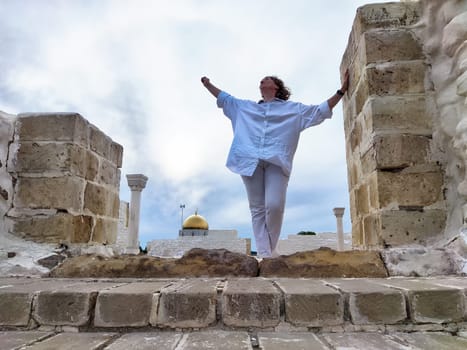 Blonde woman tourist stands confidently amidst ancient ruins, arms outstretched, embracing the adventure of exploring a historic location.