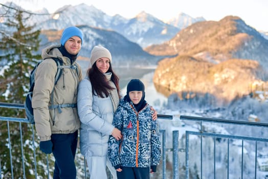 Golden Hour Family Adventure Snowy Mountain Panorama with Parents and Child Overlooking Serene Lake Landscape Capturing Warm Winter Bonding Moment in Nature - This high-resolution photograph showcases a heartwarming family scene set against a breathtaking winter mountain landscape. A family of three - father, mother, and child - stand closely together on a metal railing overlooking a panoramic vista bathed in the warm, golden light of sunset. The father, wearing a beige jacket and light gray beanie, carries a gray backpack, while the mother dons a light gray puffer jacket with a cream scarf. Their child, dressed in a blue patterned jacket with black accessories, completes the trio. Behind them, snow-covered mountain peaks reflect the golden hour glow, with a serene lake mirroring the sky's colors amidst a dense evergreen forest. This image captures the essence of family bonding, outdoor adventure, and the majestic beauty of nature, making it perfect for travel, lifestyle, and winter gear promotions.
