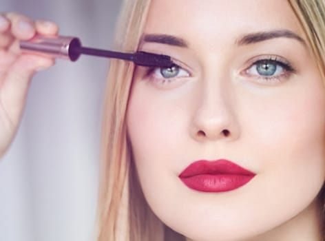 Woman applying mascara with confidence, defined brows and red lipstick. Blonde beauty focused on her eye makeup, cosmetics look