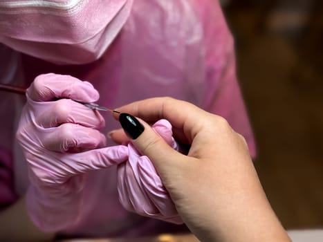Female manicurist painting black nail on caucasian young woman hand in salon.