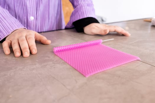 Person making pink candle with textured wax sheet on table.