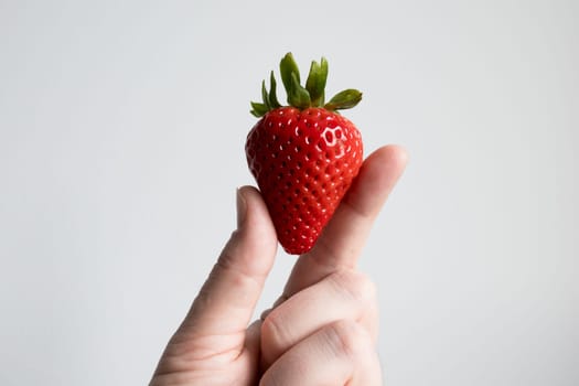 Hand holding ripe red strawberry against light background.