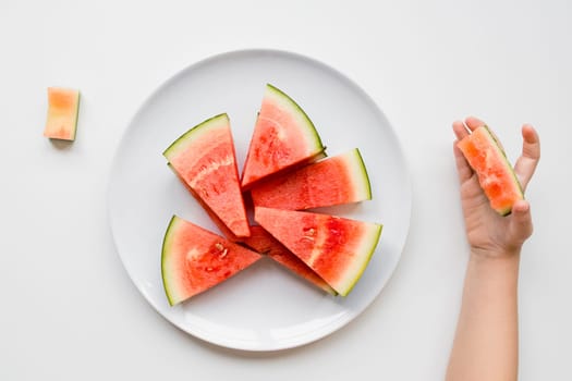 Childs hand holding watermelon slice on white plate with triangular pieces.