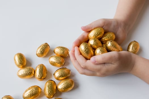 Child holding handful of golden wrapped chocolate eggs on white background.