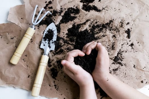 Child hands with soil and gardening tools on paper background.