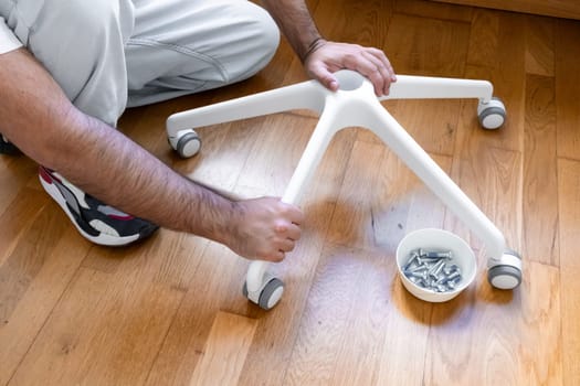 Male adult assembling office chair base with wheels on wooden floor.