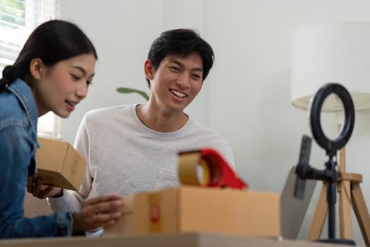 A smiling young man and woman are delightfully unboxing products while connecting with their audience through a live stream, showcasing their entrepreneurial journey.
