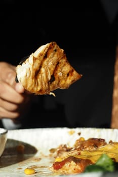 A person holds a piece of grilled chicken above a plate of flavorful food.