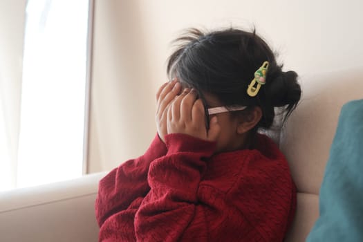 A young child is sitting on a couch, hiding her face with hands indoors.