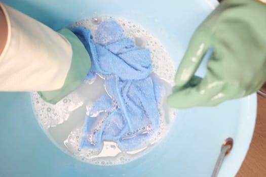 Hands in green gloves wash a blue cloth in a basin filled with bubbles.