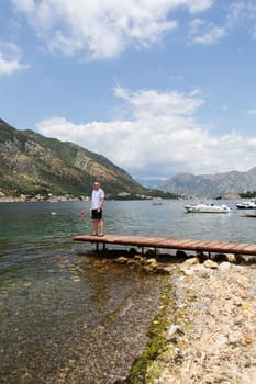 A man relaxes on a wooden dock overlooking a calm bay, mountains rising in the background on a sunny day.