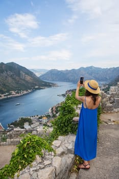 A girl in a blue dress and a straw hat stands against the backdrop of the very beautiful Bay of Kotor in Montenegro, and takes pictures with her phone