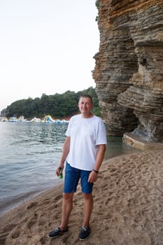 Happy man enjoying a drink on a sandy beach near a rocky cliff, sunny summer day.