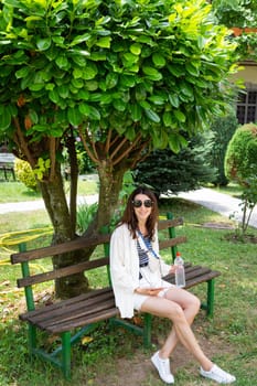 A young woman relaxes on a park bench under a lush green tree, enjoying a drink and her music.