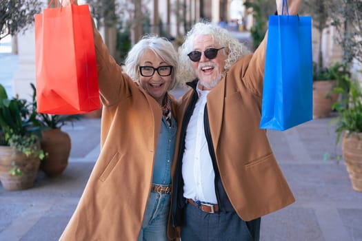 Joyful senior couple celebrates after shopping, holding colorful bags up high. Dressed in stylish coats, they smile happily while standing outdoors in a charming urban setting with plants.