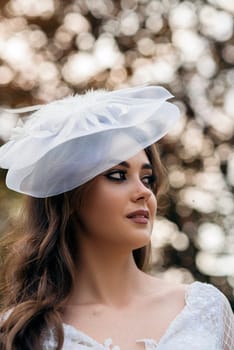 portrait of a young beautiful bride in a white dress and a white hat. white hat with a veil in a vintage style. in a spring park on a wedding day, a bright brunette in a stylish dress. against the background of bokeh