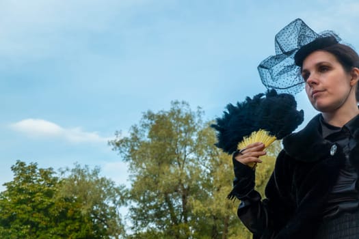 A confident and sophisticated lady in a strict black suit of the 19th century posts with a fan against the backdrop of an park.