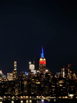 The Empire State Building glows in red, blue, and gold above the New York City skyline, reflecting in the calm nighttime river.