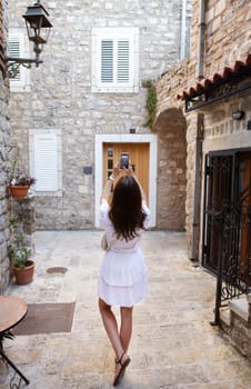 A woman in a white dress takes a photo on her phone in a quaint stone alley. Travel in Europe