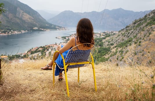 A woman in a blue dress relaxes on a yellow chair, enjoying a breathtaking view of a serene bay surrounded by mountains.