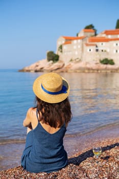 Sveti Stefan Island, Montenegro July 5, 2021: Adriatic Sea. A girl in a straw hat sits on the beach against the backdrop of St. Stephen's Island