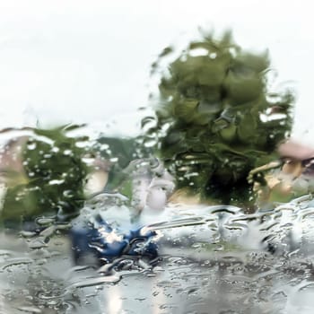 Rain shower on car windshield or car window and blurry road in background. Drizzle raining decreases driving visibility. Driving in rainy season. 
