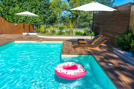 Backyard swimming pool with turquoise water, wooden deck, umbrellas, lounge chairs, and pink donut float