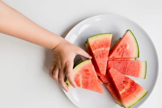 Child hand reaching for sliced watermelon on white plate.