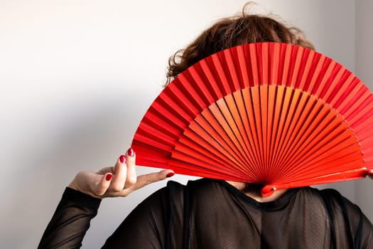 Caucasian female holding red fan covering face with stylish black top.