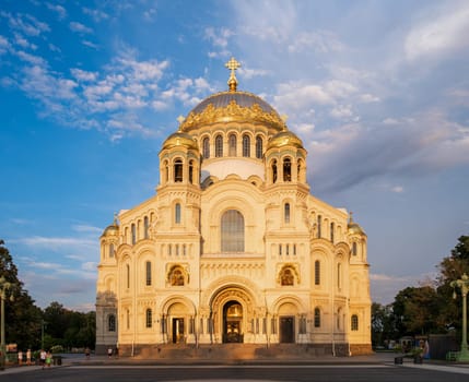 Kronstadt, Russia - 07.27.24. Naval cathedral facade illuminated by evening sunlight, showcasing intricate architectural details and golden domes against a blue sky.