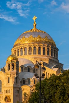 Kronstadt, Russia - 07.27.24. Naval cathedral facade illuminated by evening sunlight, showcasing intricate architectural details and golden domes against a blue sky.