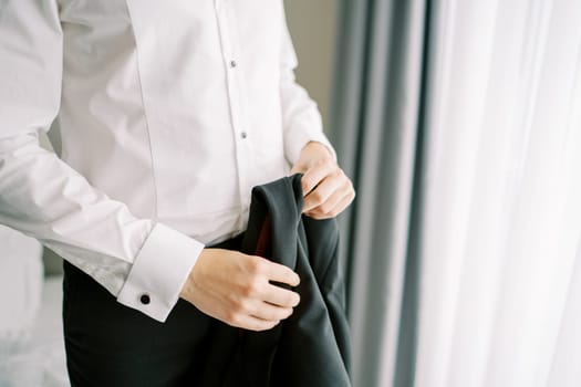 Groom holds a black jacket in his hands while standing in a hotel room. Cropped. Faceless. High quality photo