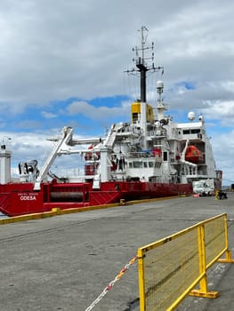Punta Arenas, Chile 10.22.2023 The white and red research vessel Odesa is moored at a concrete port, surrounded by equipment and vehicles, beneath a cloudy sky with blue patches.
