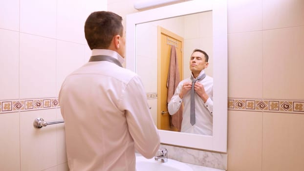 Businessman is getting ready in the morning, buttoning his shirt and adjusting his tie in front of the bathroom mirror