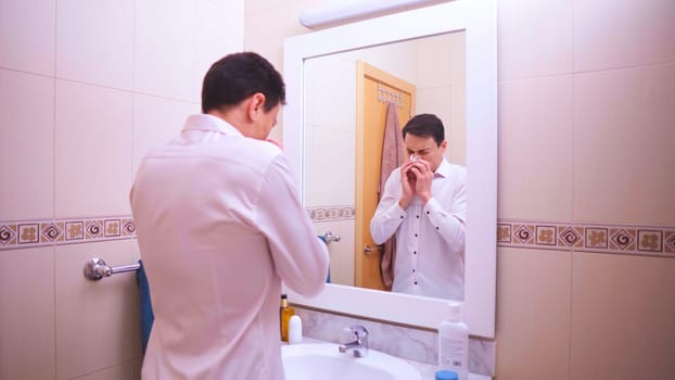 Office worker blowing his nose with a tissue while looking in the bathroom mirror