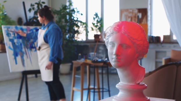 A woman paint artist drawing on a canvas with blue paint - greek head sculpture on the foreground in red neon lighting. Mid shot