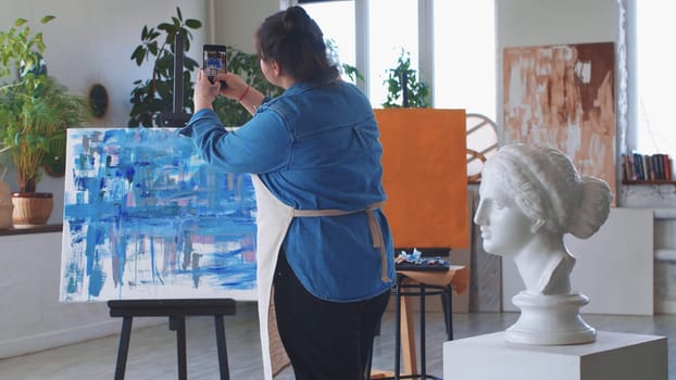 A woman paint artist taking pictures of her abstract oil painting on her phone. Mid shot