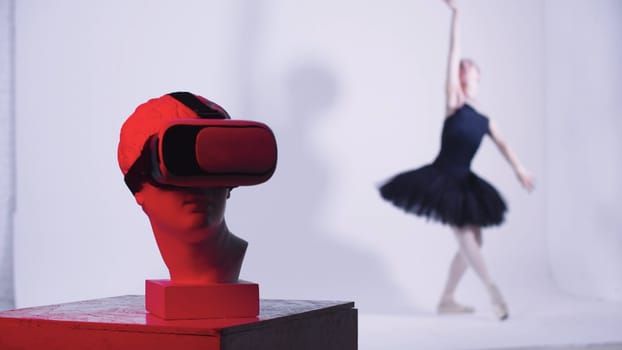 Young woman ballerina in black dress dancing - a head of greek sculpture in VR glasses on the foreground - red lighting. Mid shot
